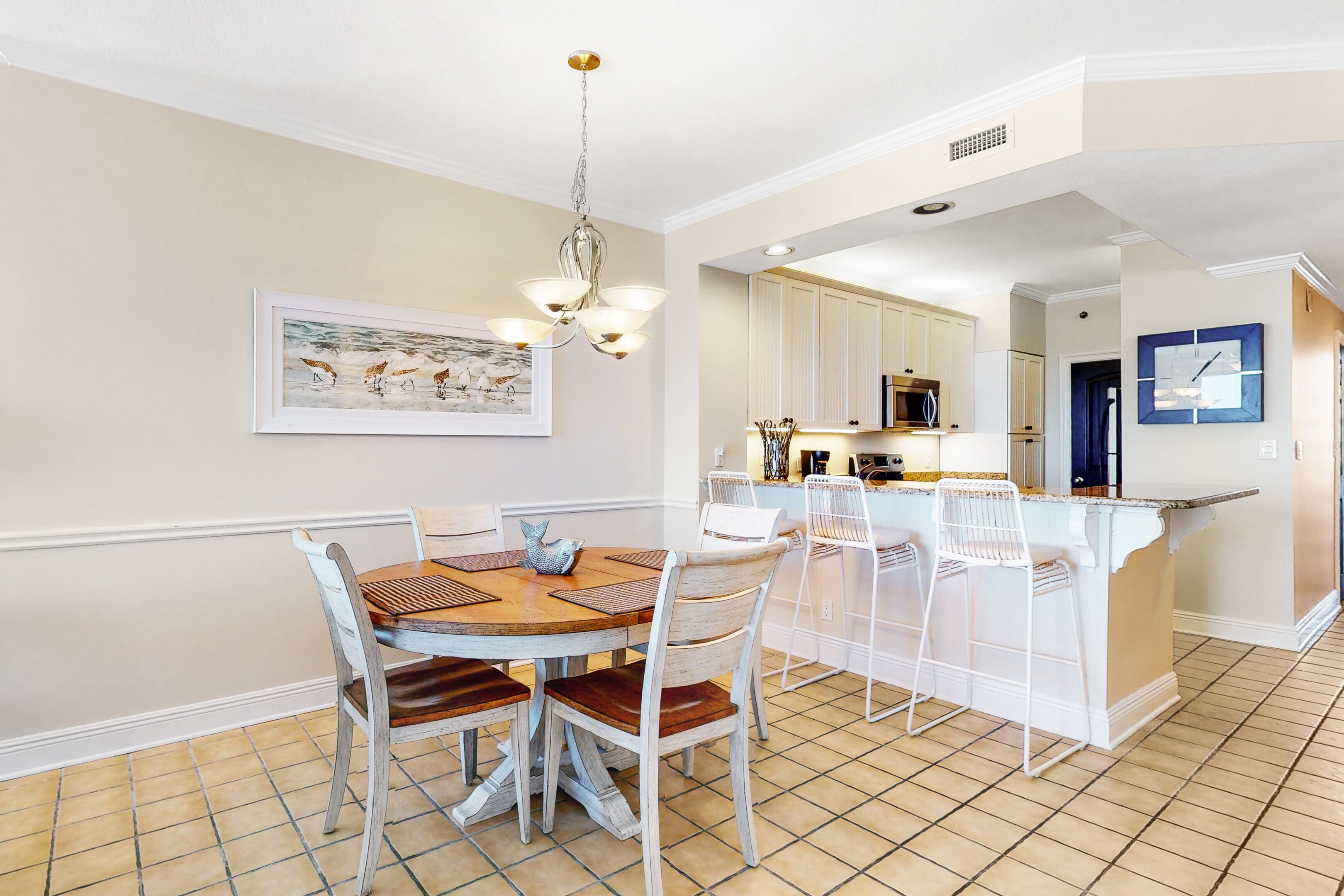 A dining area with a wooden table and chairs, a chandelier, and a kitchen in the background.