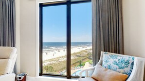 Een kamer met een groot raam dat uitkijkt op een strand, een witte stoel met kussen, en een klein tafeltje.