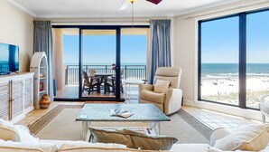Een ruime woonkamer met een groot raam dat uitzicht geeft op het strand, een plafondventilator en een televisie op een kast.