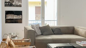 Een moderne woonkamer met een beige bank, een klein rond tafeltje en een stoel. Twee ingekaderde foto's hangen aan de muur.