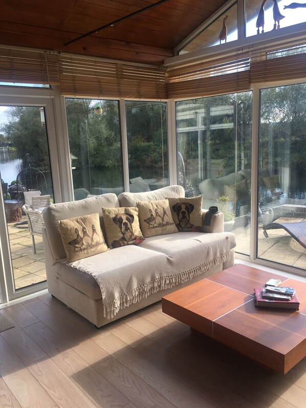 A modern living room with a white sofa, wooden coffee table, and large windows offering a view of a garden and outdoor seating area. - Riverside bungalow, Hot Tub/Sauna, Nr Legoland, Windsor, Thorpe Park Theme Park. (Wraysbury)