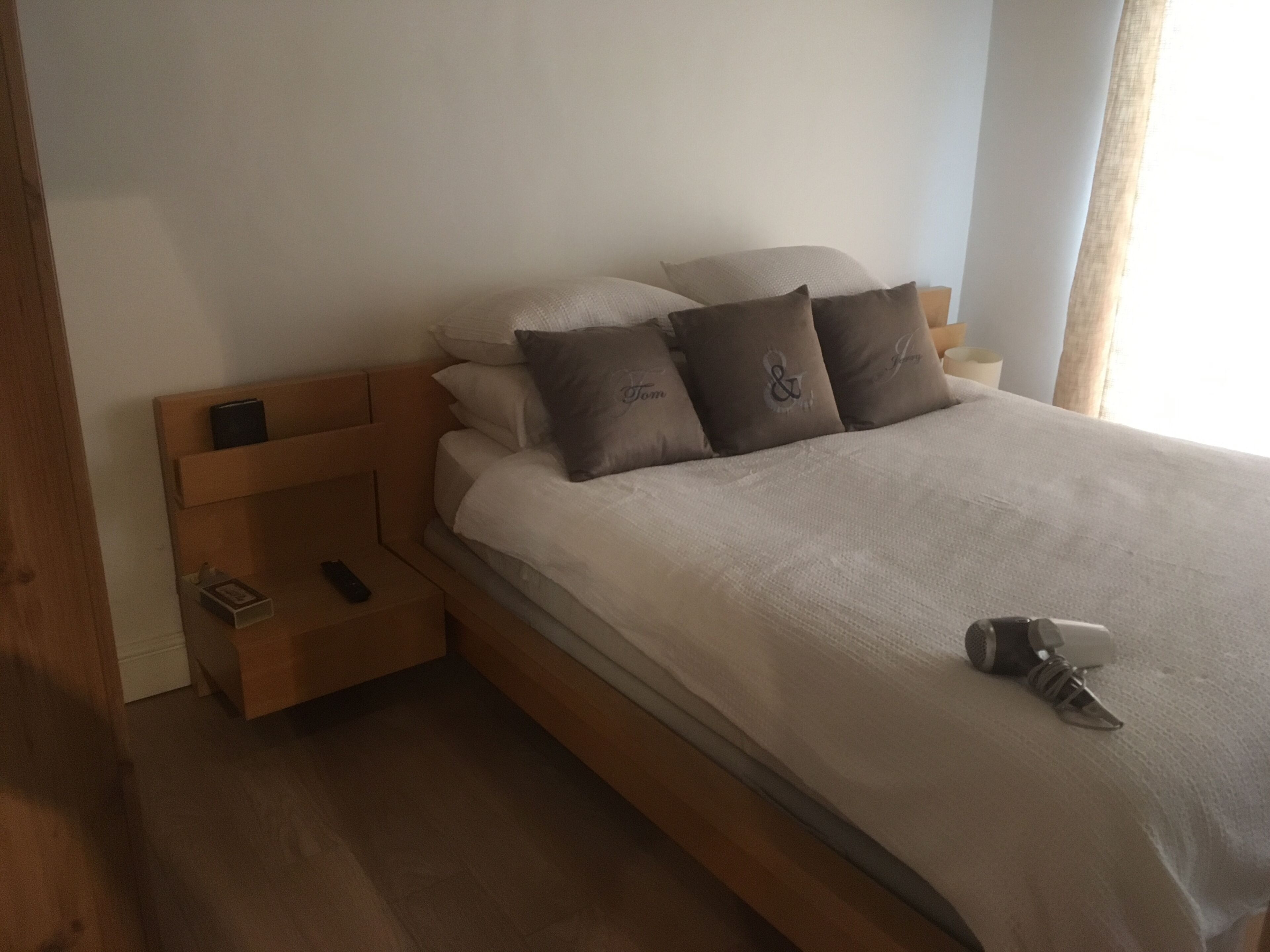 A neatly made bed with a wooden headboard, a bedside table with a remote control, and a pair of slippers on the bed.