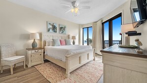 A bedroom with a bed, nightstands, a chair, a ceiling fan, artwork, and a view of the outdoors.