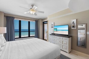 A hotel room with a bed, a television, a dresser, and a large window with a view of the beach.