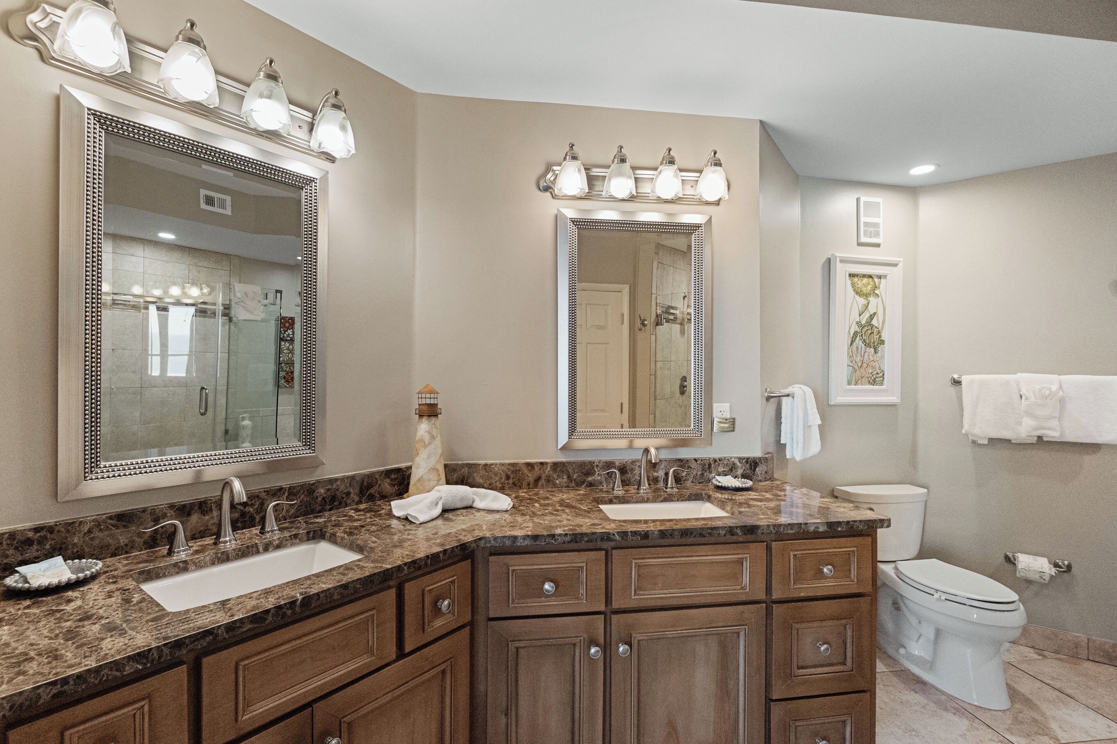 A bathroom with a marble countertop, double sinks, a large mirror with lights, wooden cabinets, a toilet, and a towel rack.