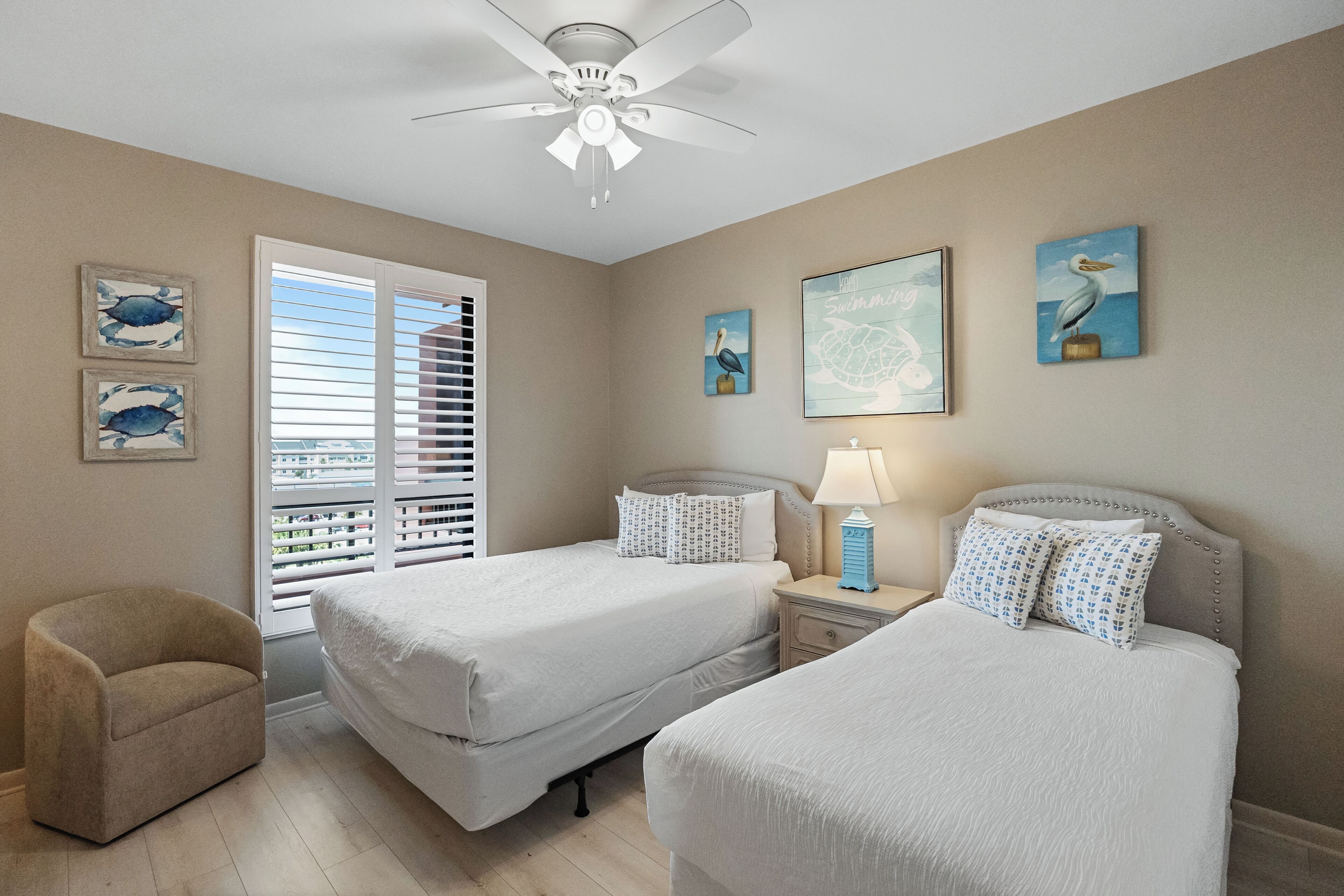 A bedroom with two beds, a ceiling fan, artwork, and a window with blinds.