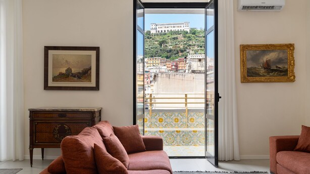 Ein Wohnzimmer mit einer Couch, einem kleinen Tisch und Blick auf ein Gebäude durch eine offene Tür.