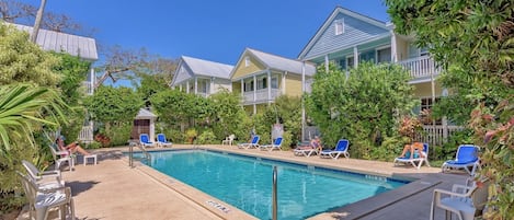 Un espace piscine doté de chaises longues et d’une piscine à l’eau d’un bleu limpide, entouré d’une végétation luxuriante et bordé d’un bâtiment avec un balcon.