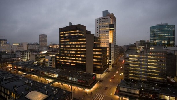 A cityscape at dusk with modern buildings, including a prominent office building with a unique corner design and a parking structure.