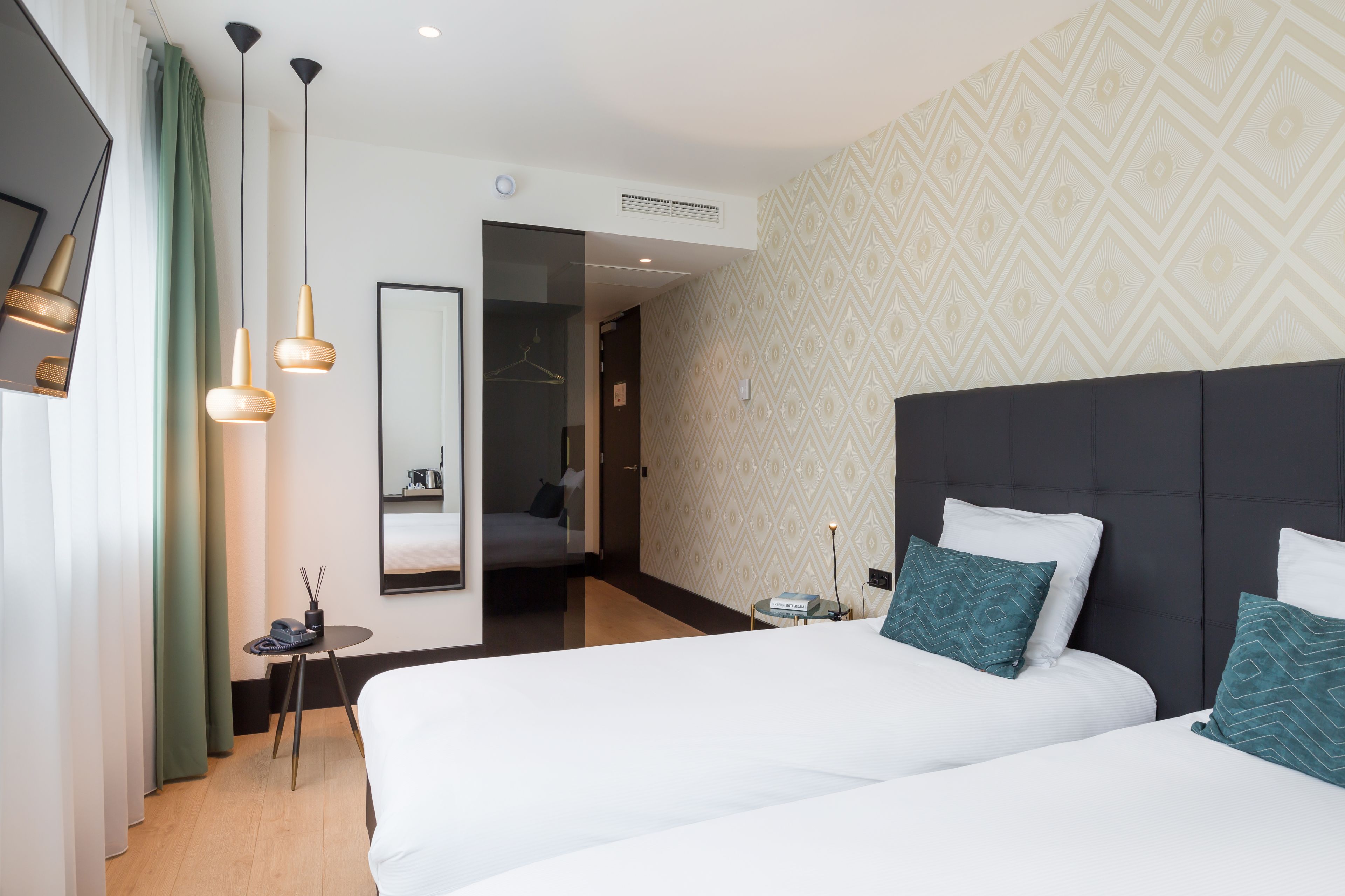 Une chambre d’hôtel avec deux lits, une petite table avec un téléphone et une tasse, un miroir et un mur décoré de motifs.