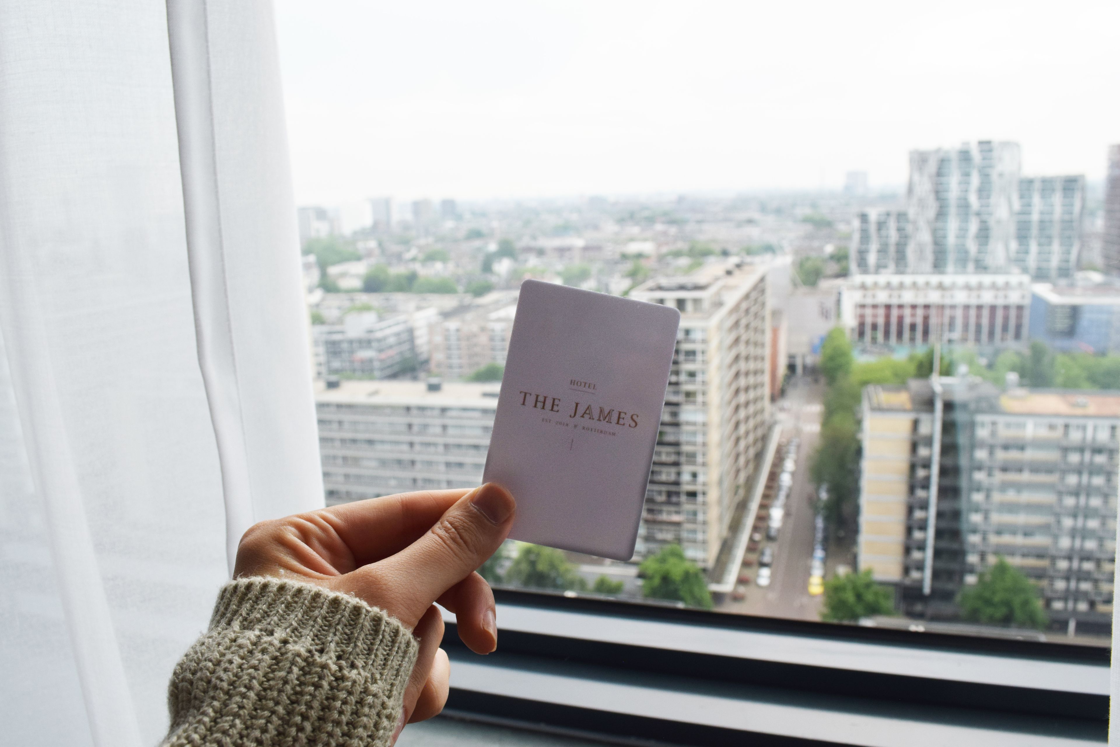 Une main tenant une carte d’hôtel sur laquelle on peut lire « THE JAMES », avec en arrière-plan une vue sur la ville à travers une fenêtre.