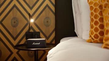 Una cama con un almohadón blanco y una manta con estampado de jirafa, un altavoz Marshall sobre una mesita y una pared decorada al fondo.