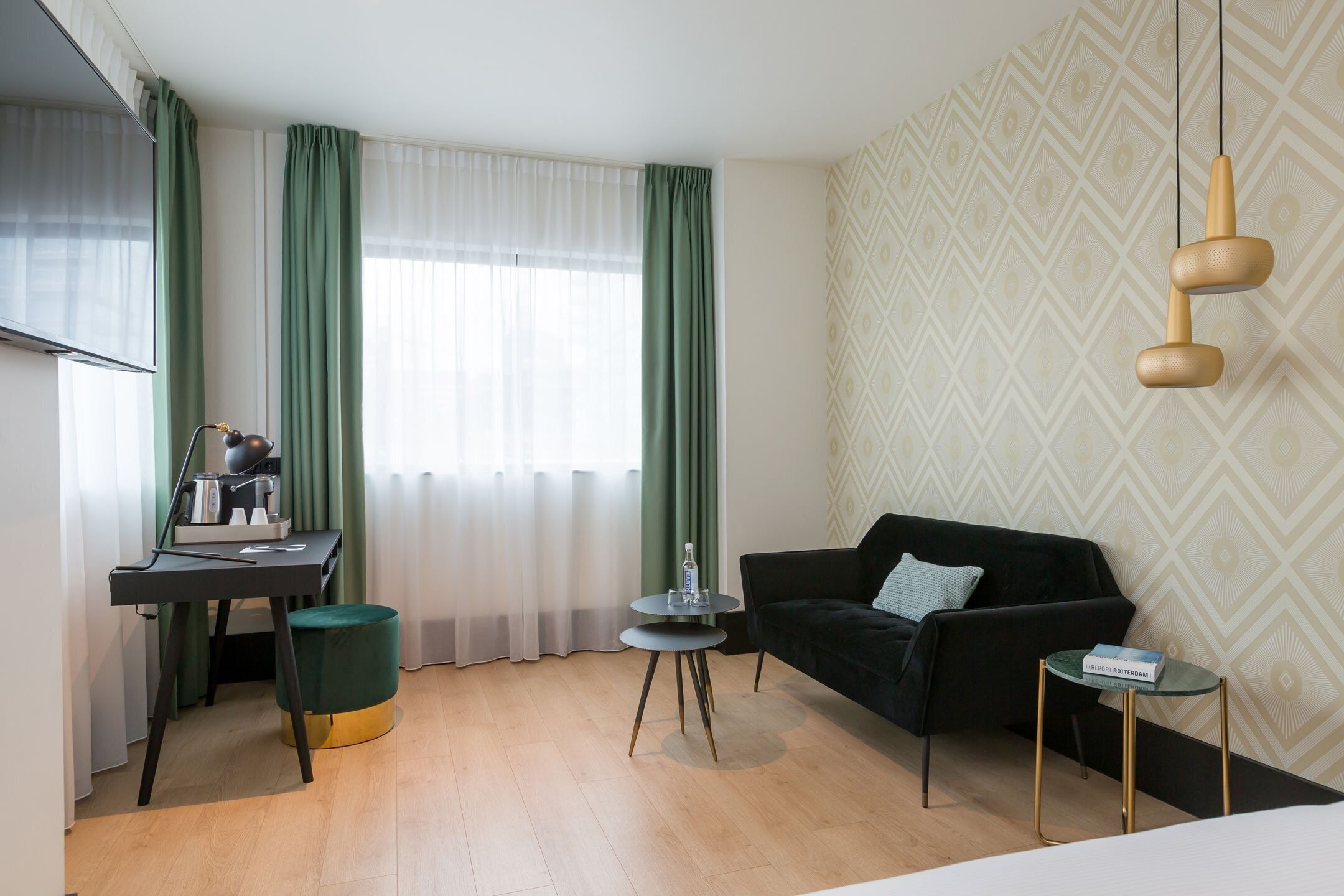 Quarto de hotel moderno com escrivaninha, sofá preto, mesa redonda pequena e luminária suspensa.