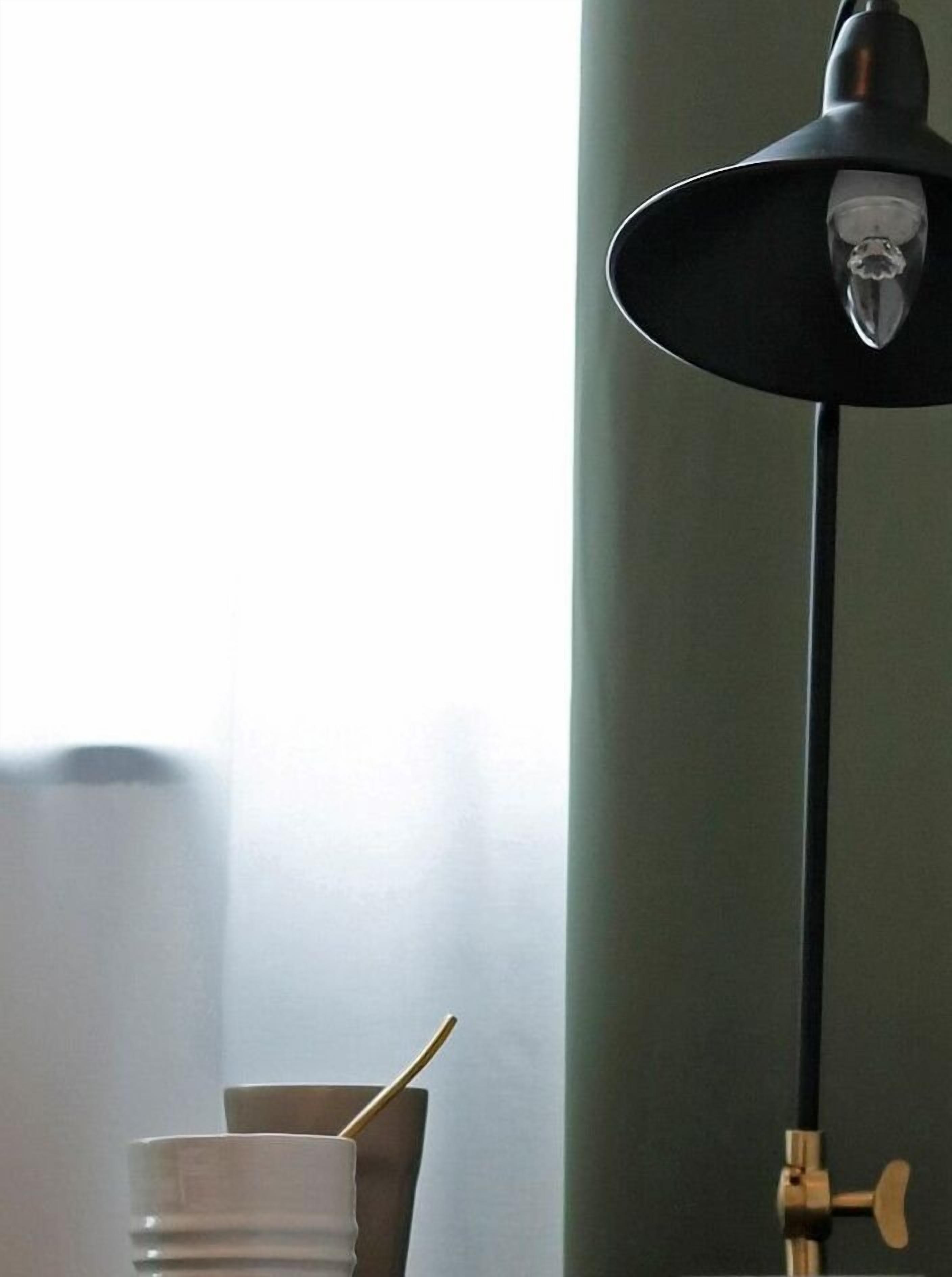 Una habitación con una lámpara de pie negra, un jarrón blanco con una cuchara dorada, y una ventana que ofrece vistas al exterior.