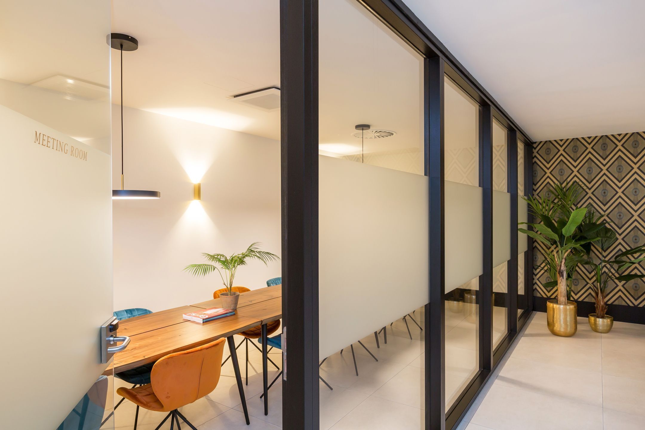 A modern meeting room with a wooden table, orange chairs, and potted plants.