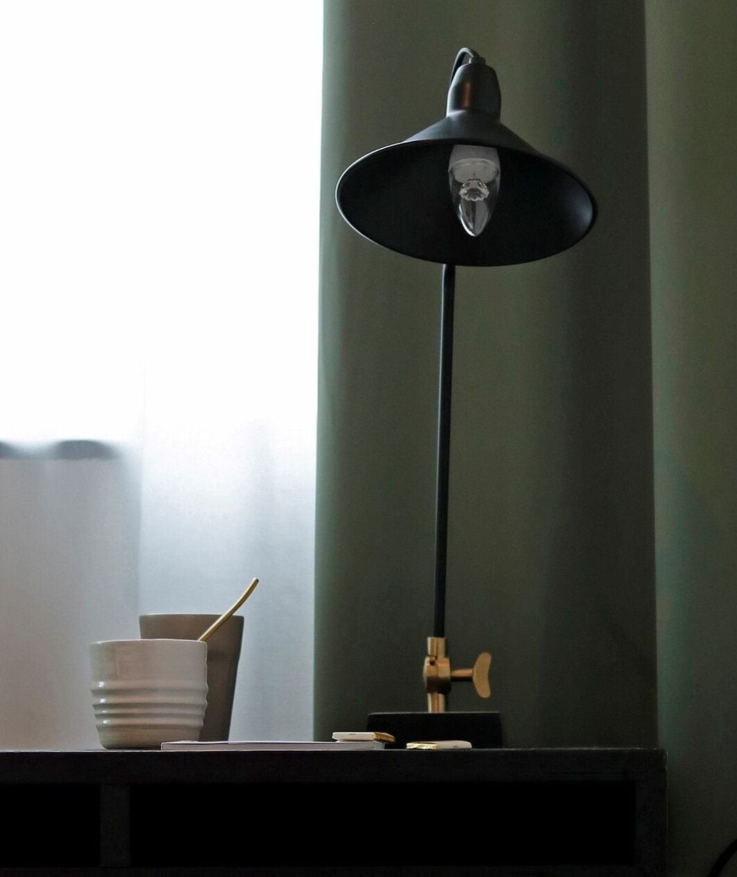 Una lámpara de escritorio con pantalla negra y bombilla visible, una taza blanca con asa dorada y un pequeño objeto de latón sobre una superficie oscura.