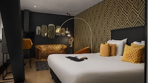 A modern hotel room with a large bed, a gold armchair, a small round table, and a patterned wall.