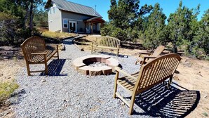 A rustic outdoor seating area with wooden chairs and a fire pit, surrounded by gravel and trees, with a house in the background. - The Cedars Country Cottage (Blanding)