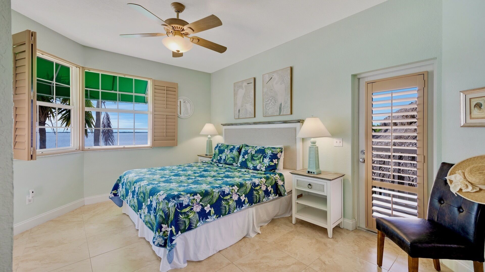Une chambre avec un lit, deux tables de chevet, un ventilateur de plafond, une fenêtre avec des volets et une porte donnant sur la plage.