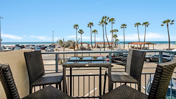 Un balcón con sillas de mimbre y una mesa con vistas a una playa con palmeras y coches aparcados.