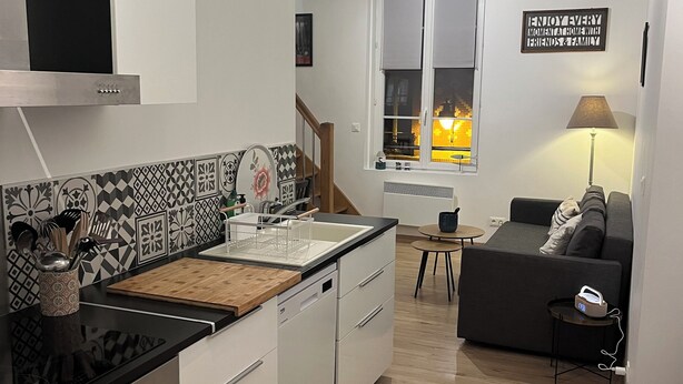 Una cucina moderna con mobili bianchi, un piano di lavoro nero e un tagliere di legno. Si vede una finestra con vista su un tramonto, un divano e una lampada nella zona giorno.