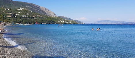 A clear beach with pebbles, calm water, and a distant mountain.