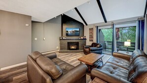 Un salon moderne avec des canapés en cuir, une cheminée, un téléviseur à écran plat et une grande fenêtre donnant sur l’extérieur.