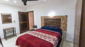 A bedroom with a bed, a ceiling fan, a door, a small table, and a wall-mounted shelf.