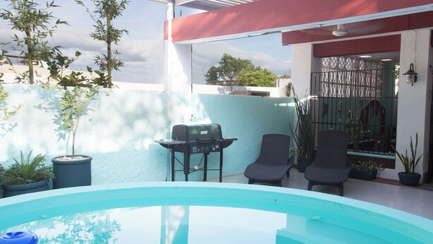 Una piscina con una parrilla y sillas, rodeada de plantas en macetas y una estructura con techo rojo.