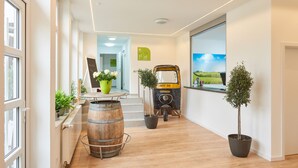Un moderno vestíbulo de hotel con piso de madera, plantas en macetas, un carrito antiguo y un amplio ventanal con vista al campo.