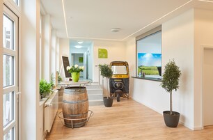 Un moderno vestíbulo de hotel con piso de madera, plantas en macetas, un carrito antiguo y un amplio ventanal con vista al campo.