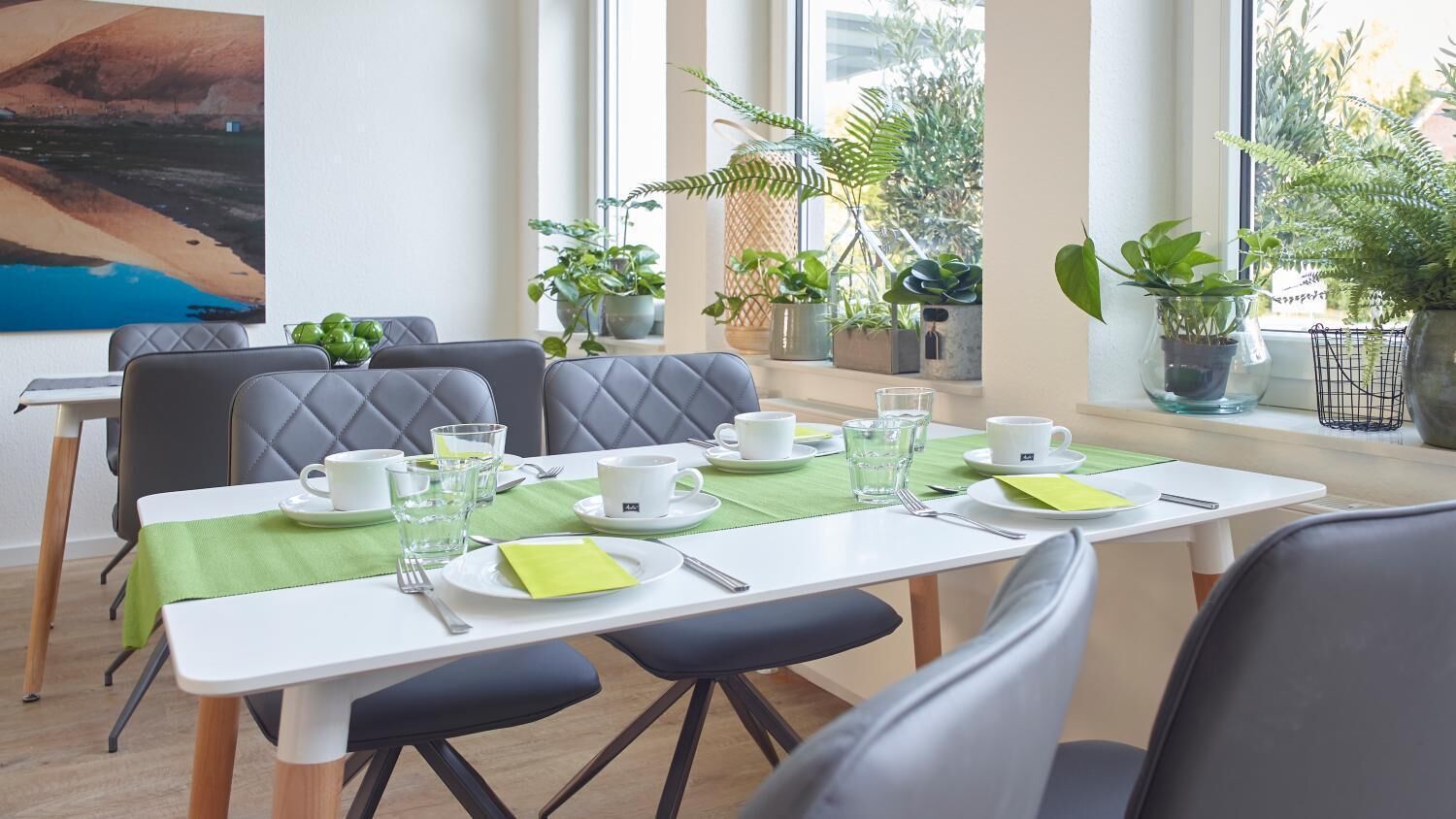 A dining area with a table set for four, surrounded by chairs, with potted plants and a painting on the wall.