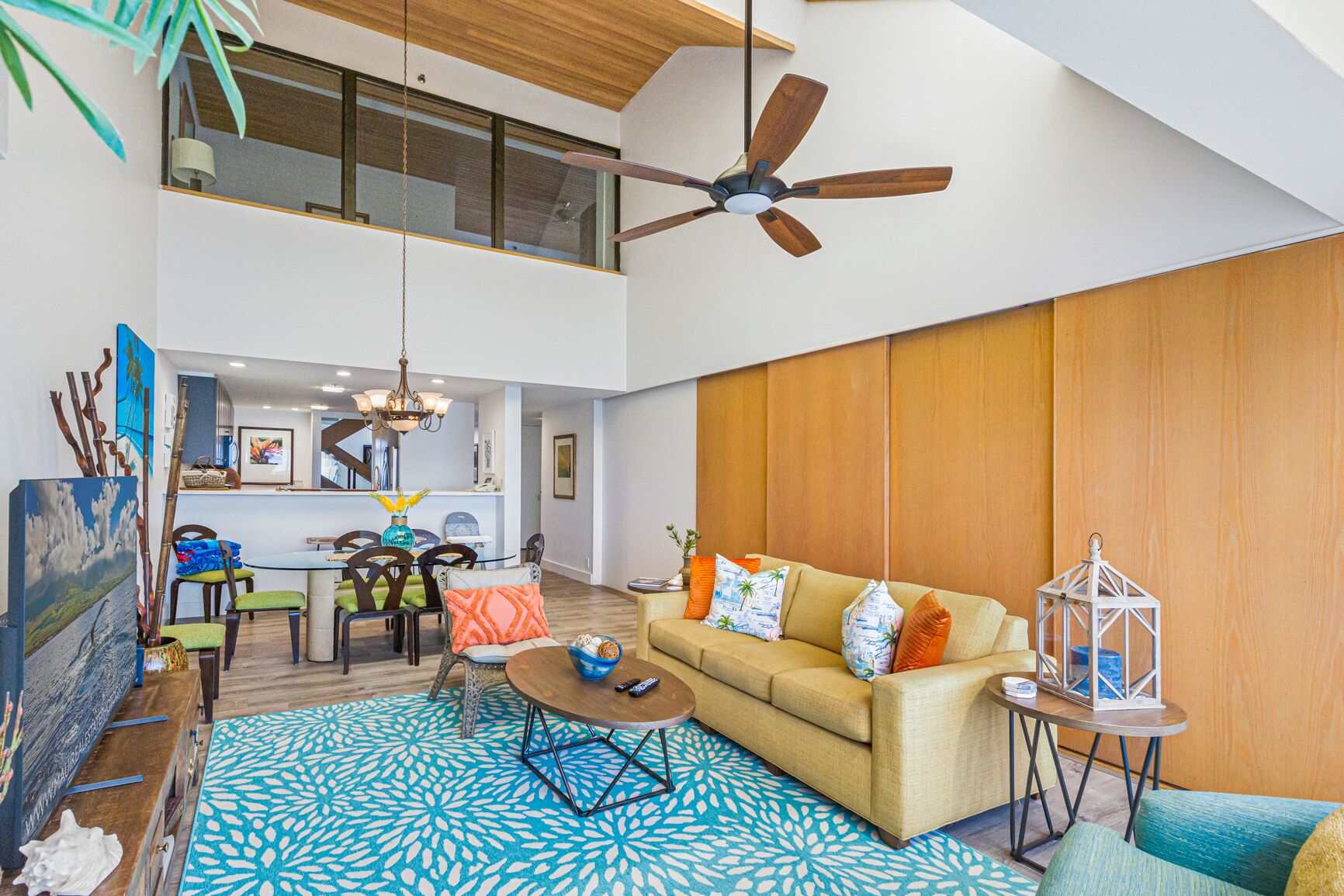 A spacious living area with a high ceiling fan, a sofa with colorful cushions, a dining table with chairs, and a patterned rug.