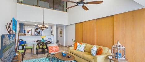 A spacious living area with a high ceiling fan, a sofa with colorful cushions, a dining table with chairs, and a patterned rug.