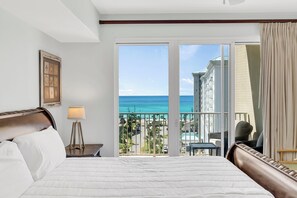 A hotel room with a large bed, a nightstand, a lamp, and a view of the ocean through a sliding glass door.