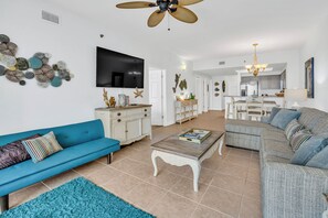 A spacious living area with a blue sofa, a white cabinet, a television, and a dining area with a chandelier. - Fabulous 2 BR condo! 2 Huge Pools! Fantastic Ocean Views! Great Location! (Miramar Beach)