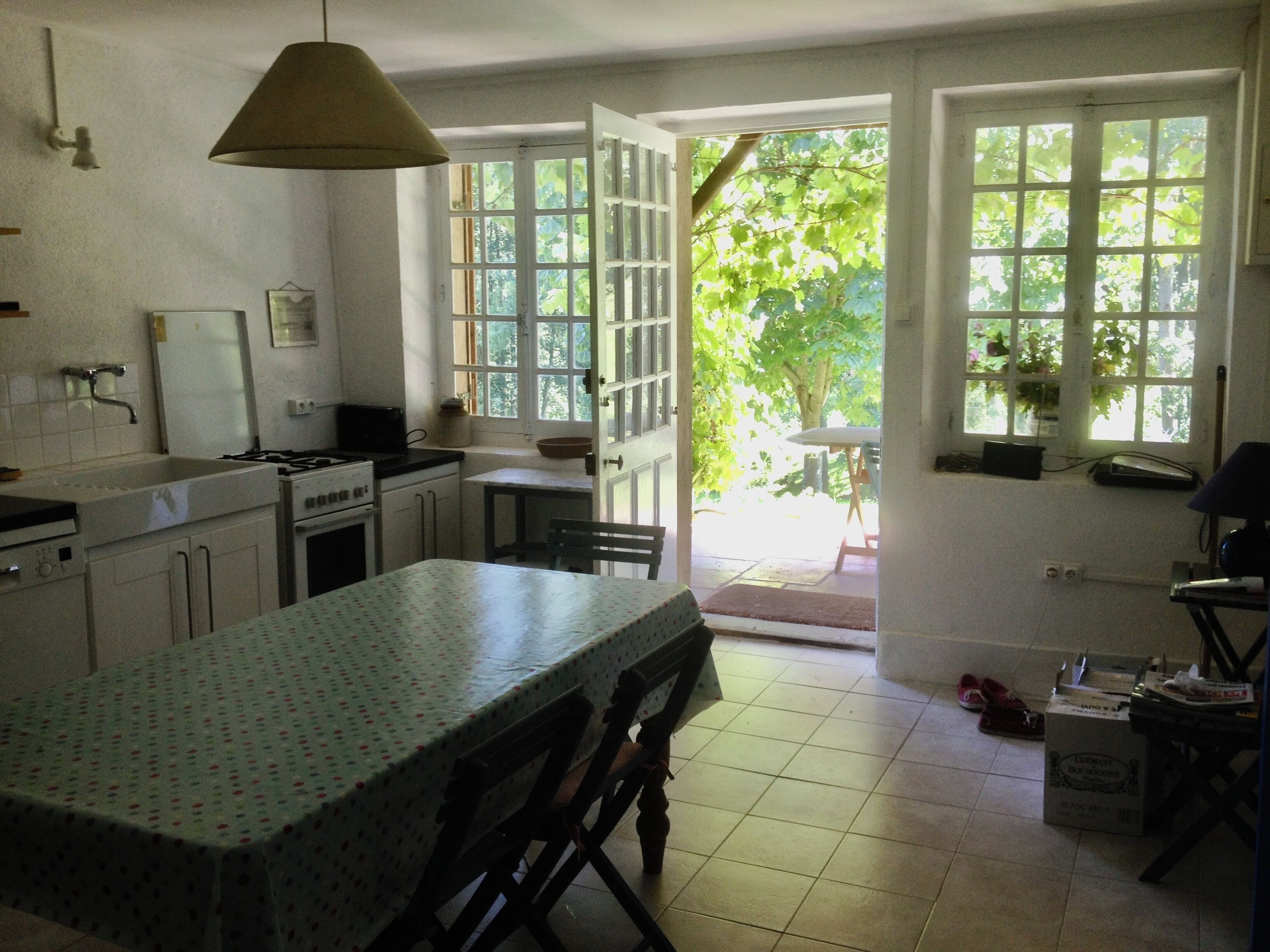 Une cuisine avec des meubles blancs, un fourneau et une table à manger recouverte d’une nappe à pois. Une fenêtre offre une vue sur la verdure à l’extérieur.