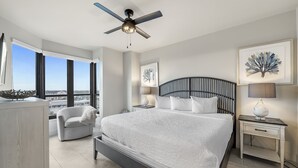 Une chambre dotée d’un grand lit, d’un ventilateur de plafond, d’une table de chevet avec une lampe, d’une chaise et offrant une vue sur la ville grâce à de grandes fenêtres.