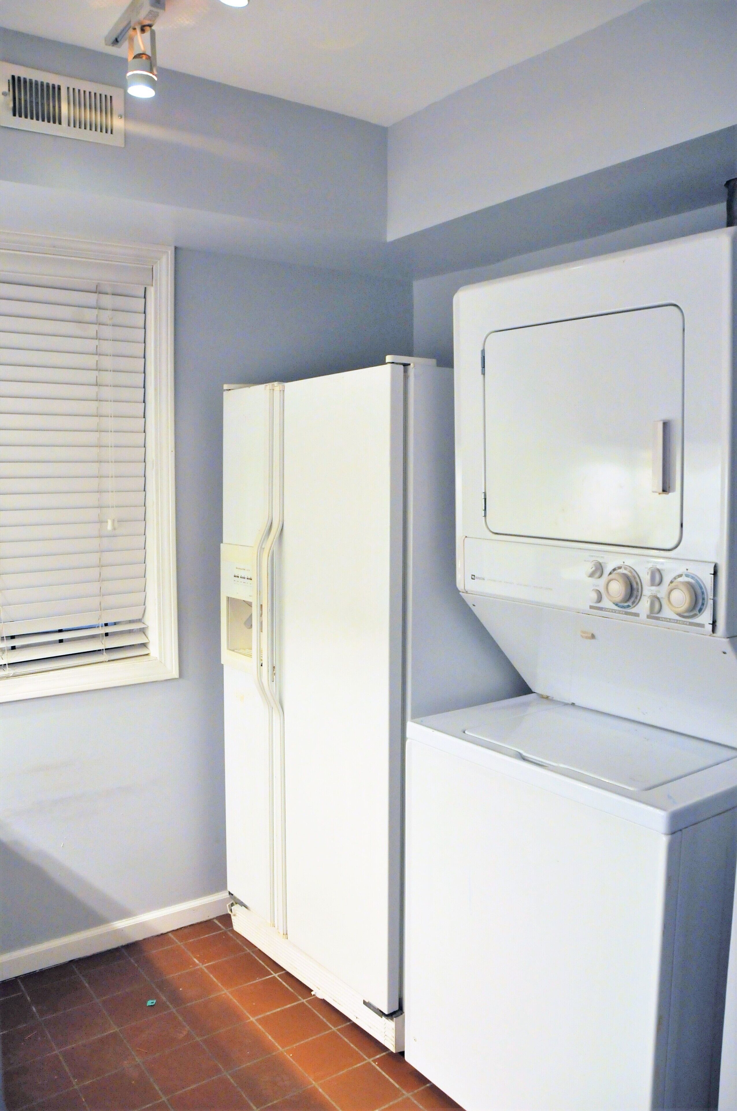 A compact laundry room with a white refrigerator and washer, a window with blinds, and a vent on the ceiling.