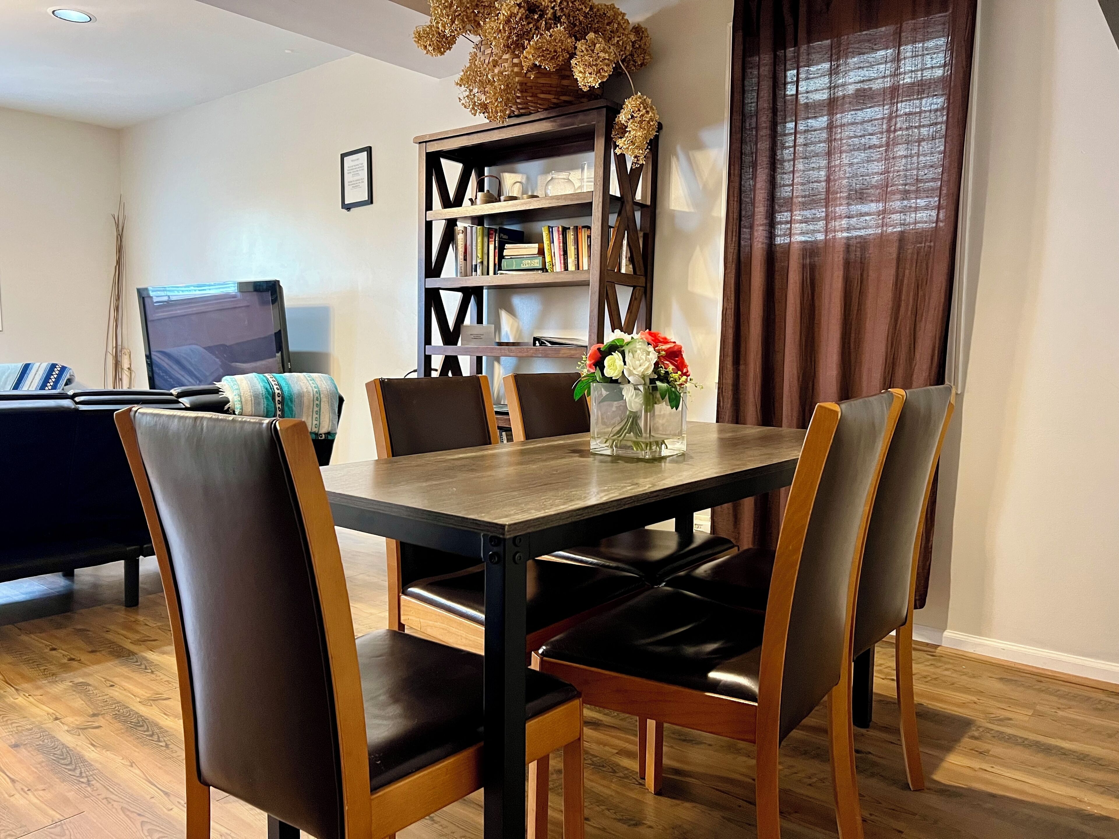 Een eethoek met een houten tafel en stoelen, een boekenkast en een televisie op de achtergrond.