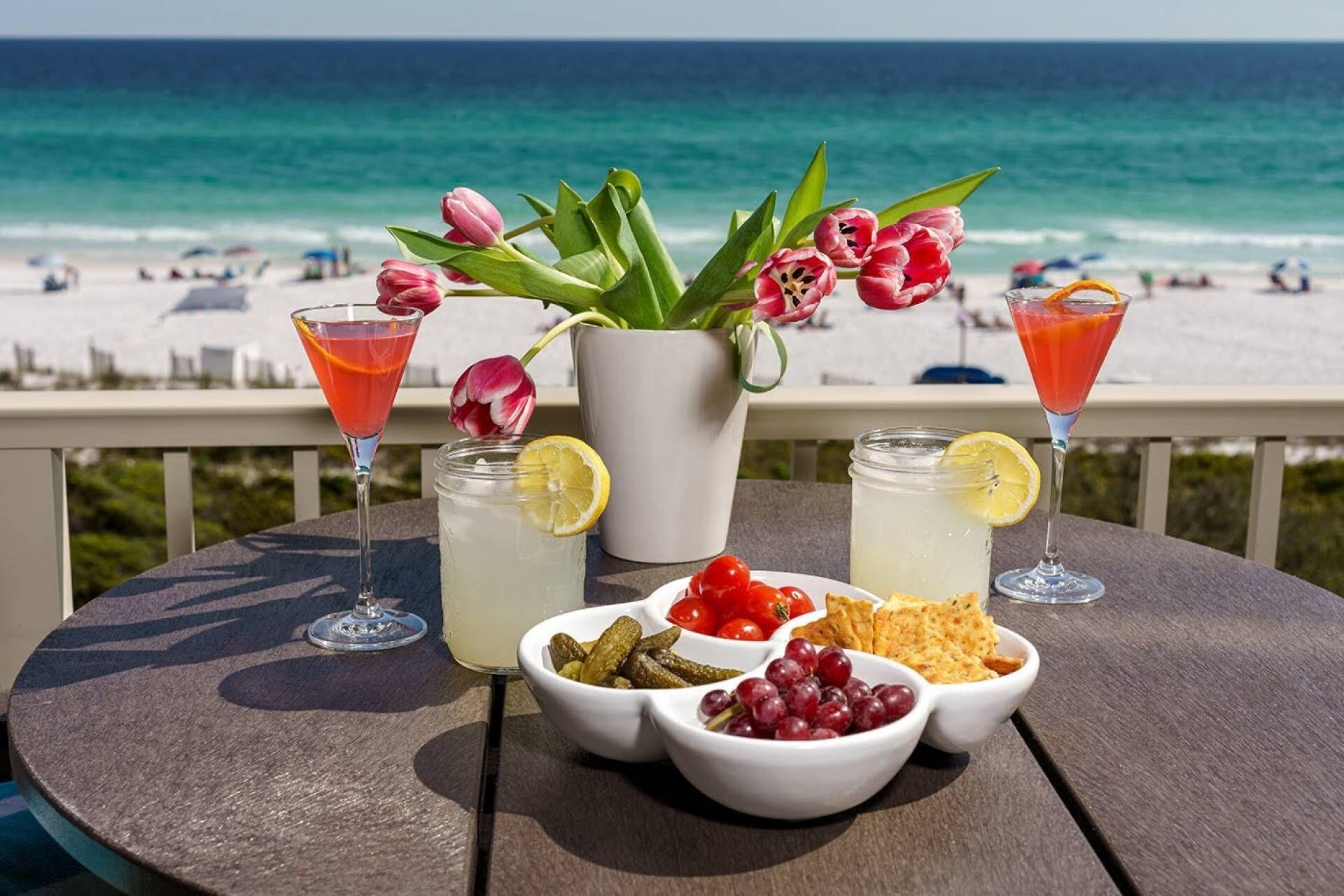 A table with drinks, a pitcher of lemonade, and bowls of snacks on a balcony overlooking a beach.