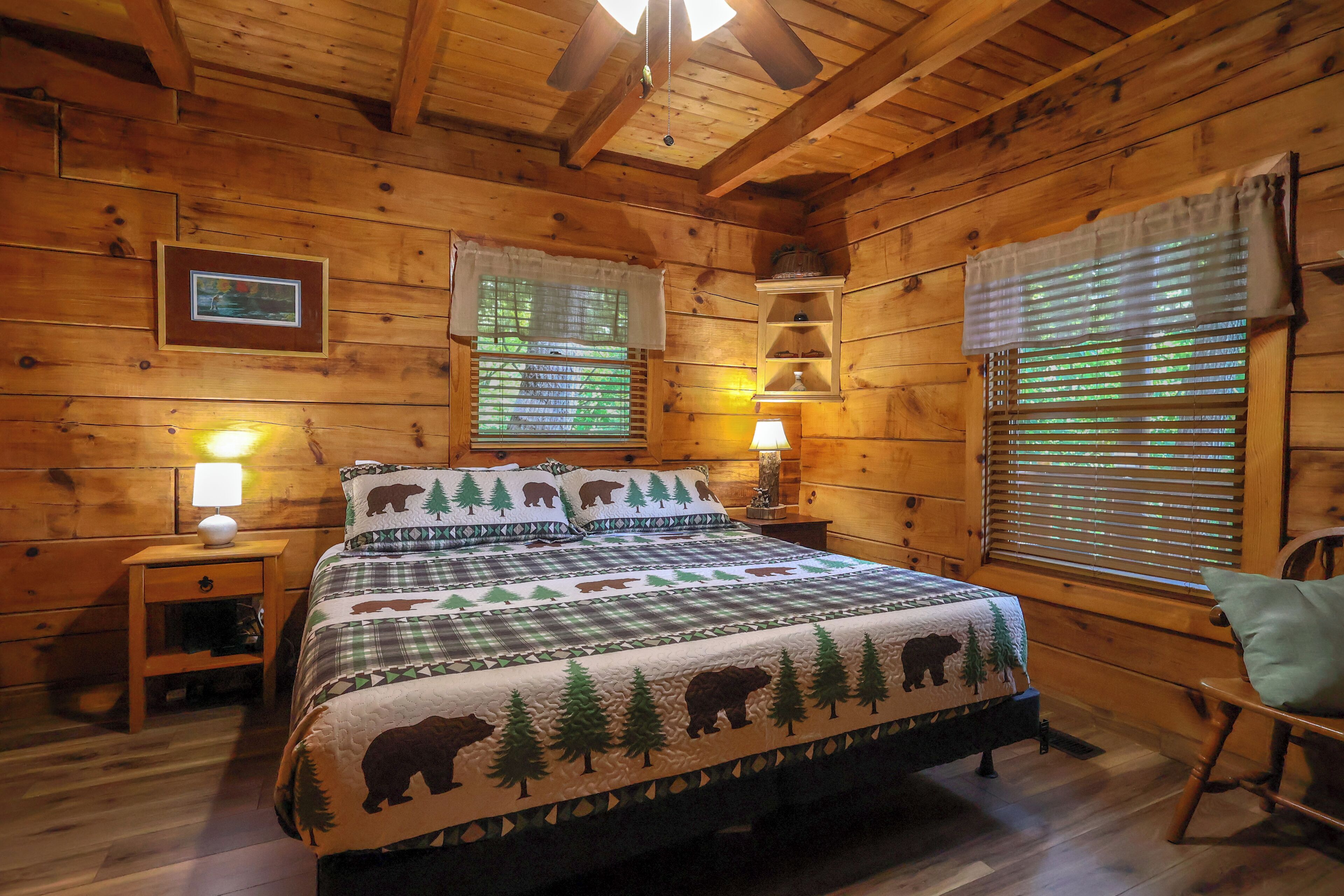 A wooden cabin bedroom with a bed, two bedside tables, a chair, a window with blinds, and a ceiling fan.