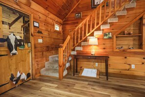 Interior de una cabaña de madera con escalera, una puerta donde se cuelgan abrigos y una mesa con lámpara.