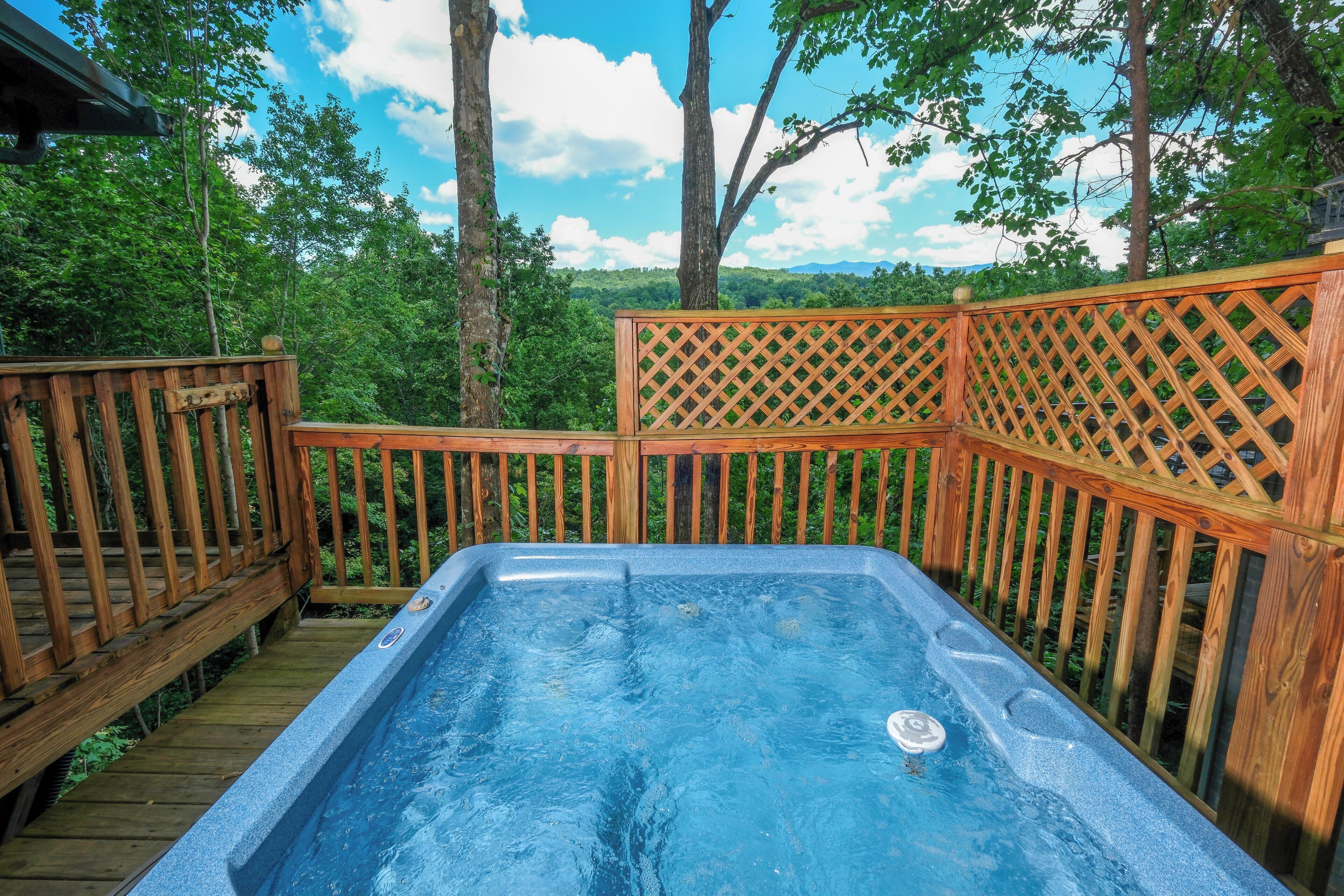 A hot tub on a wooden deck with a lattice railing, surrounded by trees and a clear sky.