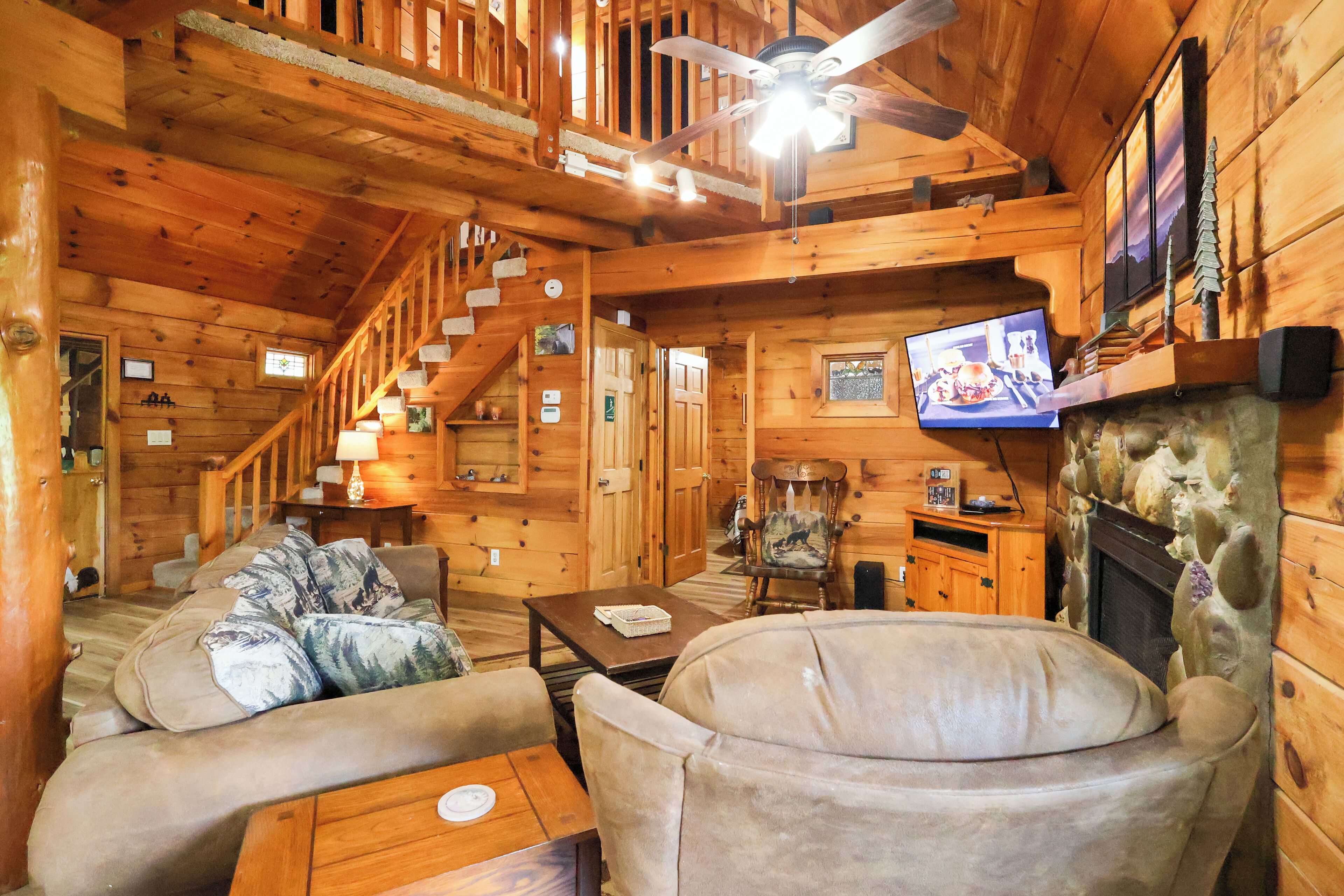 A log cabin interior with a sofa, armchairs, a fireplace, a television, and a staircase.