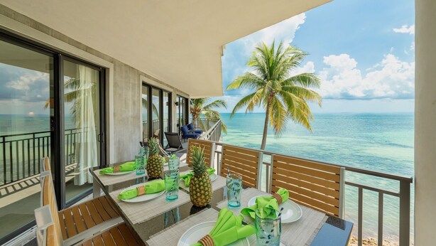 Un balcon avec une table dressée pour deux personnes, donnant sur une plage aux eaux turquoise et limpides, bordée de palmiers.