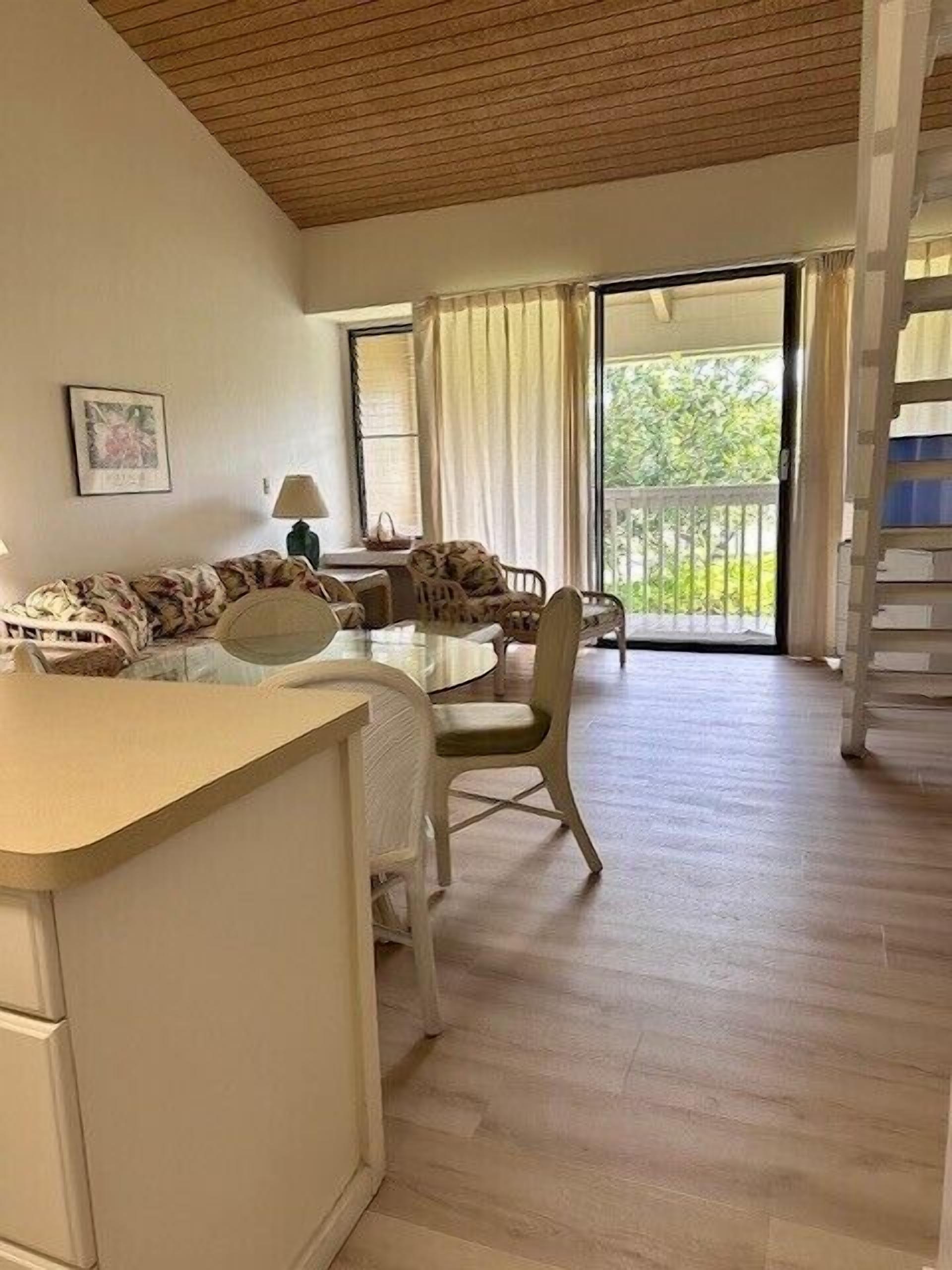 A room with a dining table and chairs, a sofa, a lamp, and a view of greenery through the window.