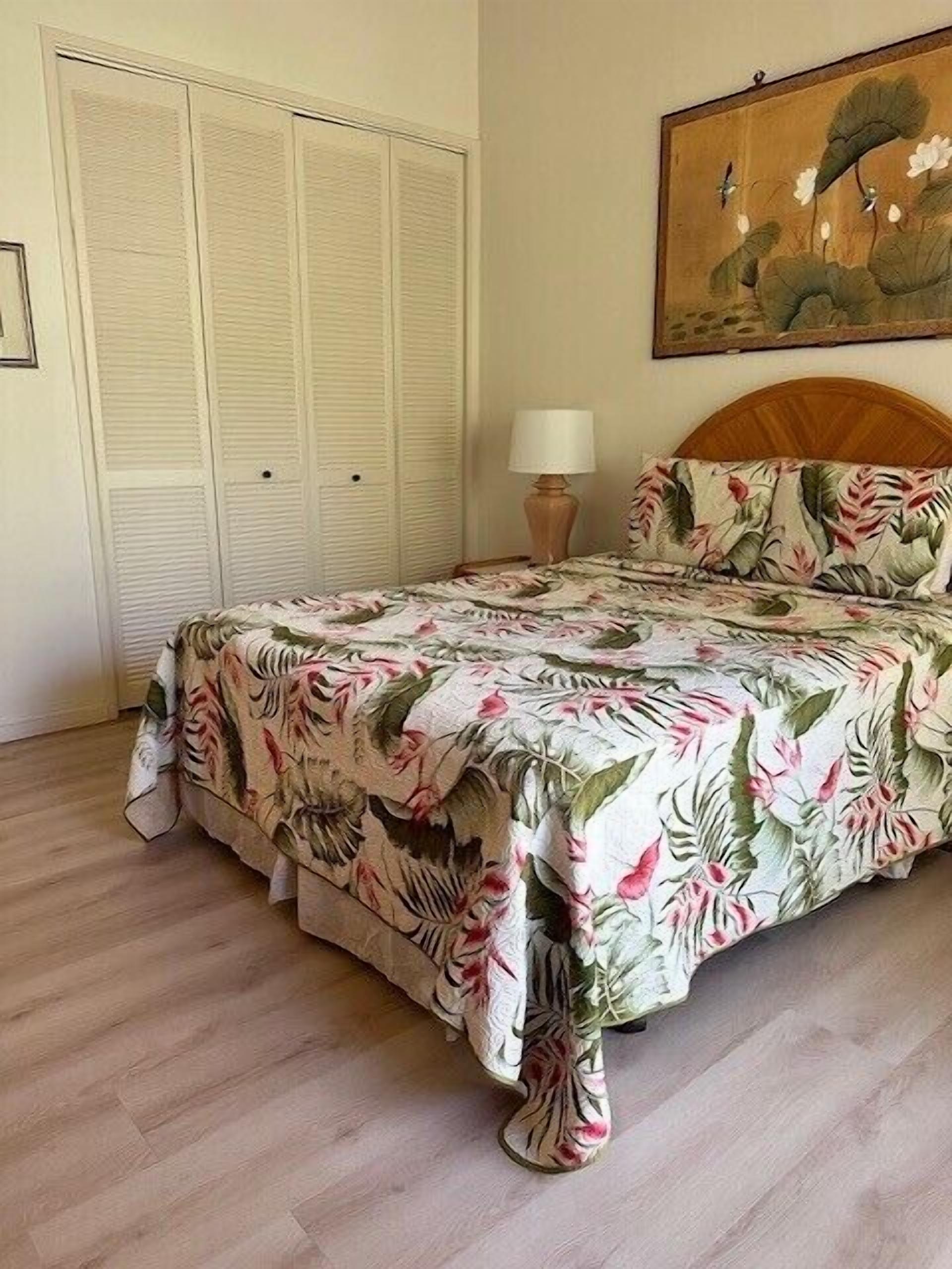 A bedroom with a bed, a nightstand with a lamp, a wooden headboard, a painting on the wall, and a set of white closets.