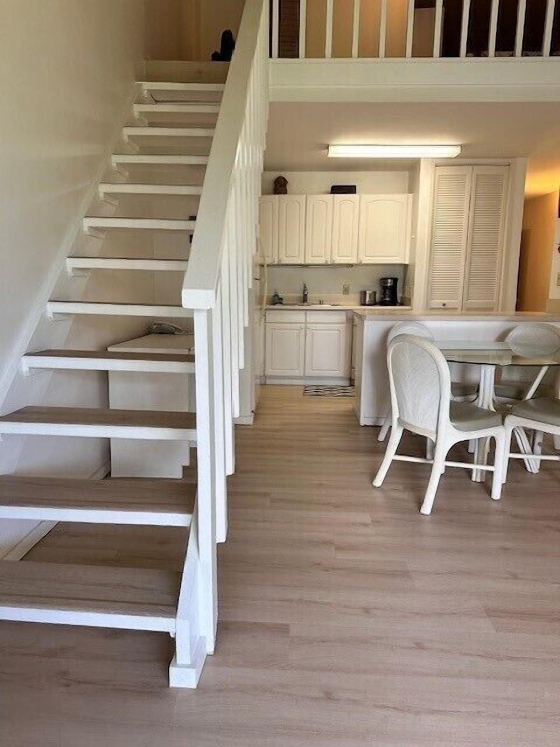 Una escalera conduce a una cocina con armarios blancos y un comedor con una mesa y sillas.
