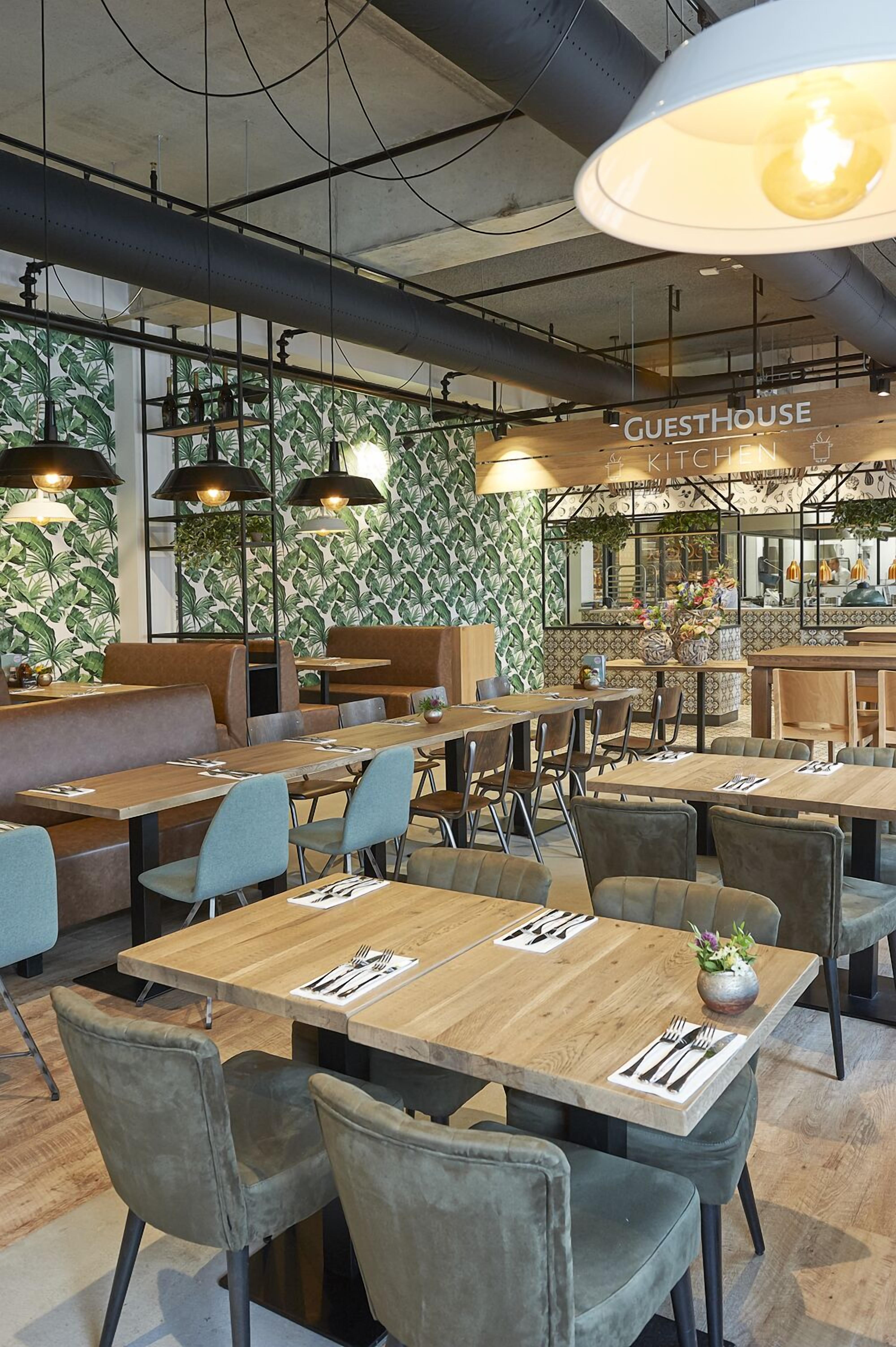 Interior moderno de restaurante con mesas y sillas de madera, lámparas colgantes negras y un diseño de papel tapiz con hojas verdes.